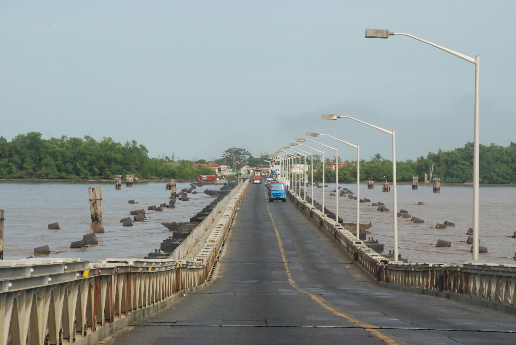 Road bridge with a row of lamp posts crossing a wide brown river in Guyana, with trees on the far bank
